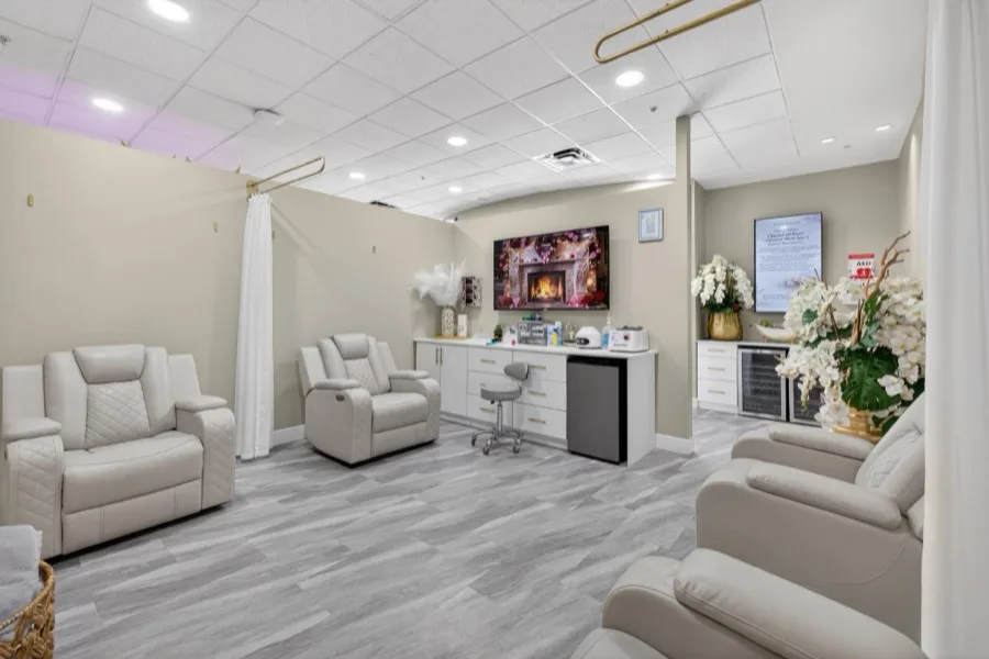 A room of cream leather IV recliners facing a wall screen, with white orchids, treatment cabinets and a glass-fronted fridge