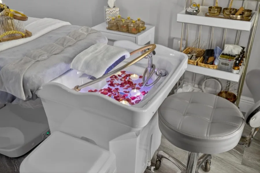 A white head-spa basin filled with red rose petals, gold fittings and a round stool beside shelves of tools