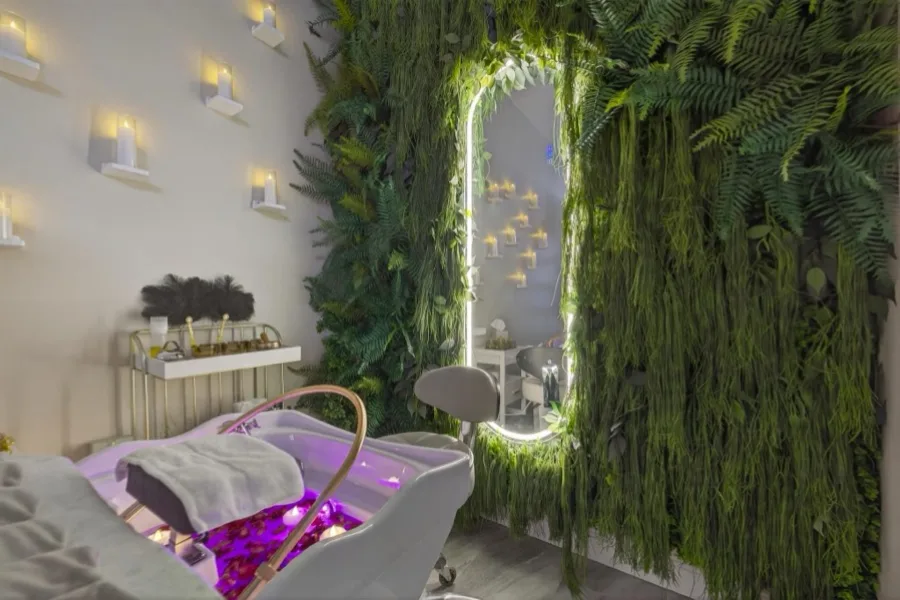 A head-spa bed under an illuminated arched mirror set into a dense fern wall, lit by candle sconces along the side wall