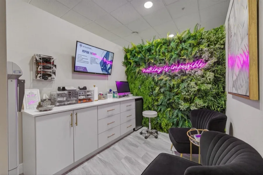 A treatment room wall of preserved ferns with a pink neon script reading nothing is impossible, beside white cabinets of products and a wall-mounted screen