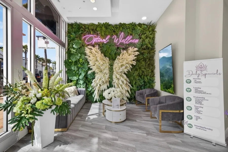 A green living wall with pink neon Optimal Wellness script between two tall white pampas wing installations, with velvet chairs and floor-to-ceiling windows