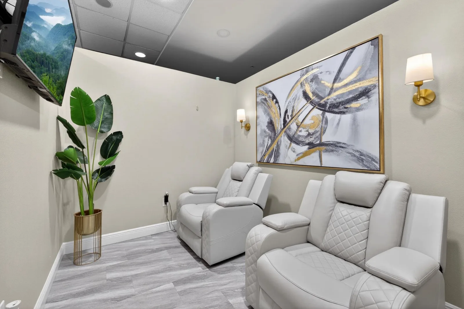 Two white quilted recliners in a private treatment room with gold-framed abstract art and wall sconces