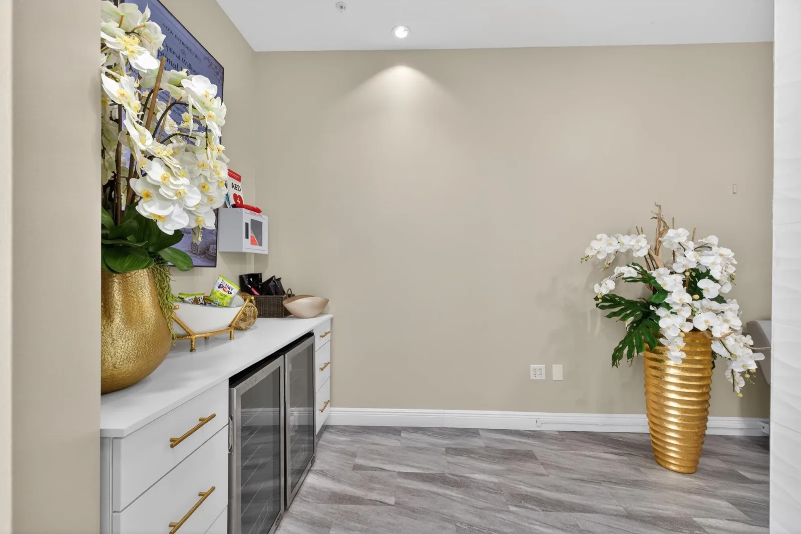 A white counter with gold vases of white orchids and a glass-fronted drinks fridge below wall screens