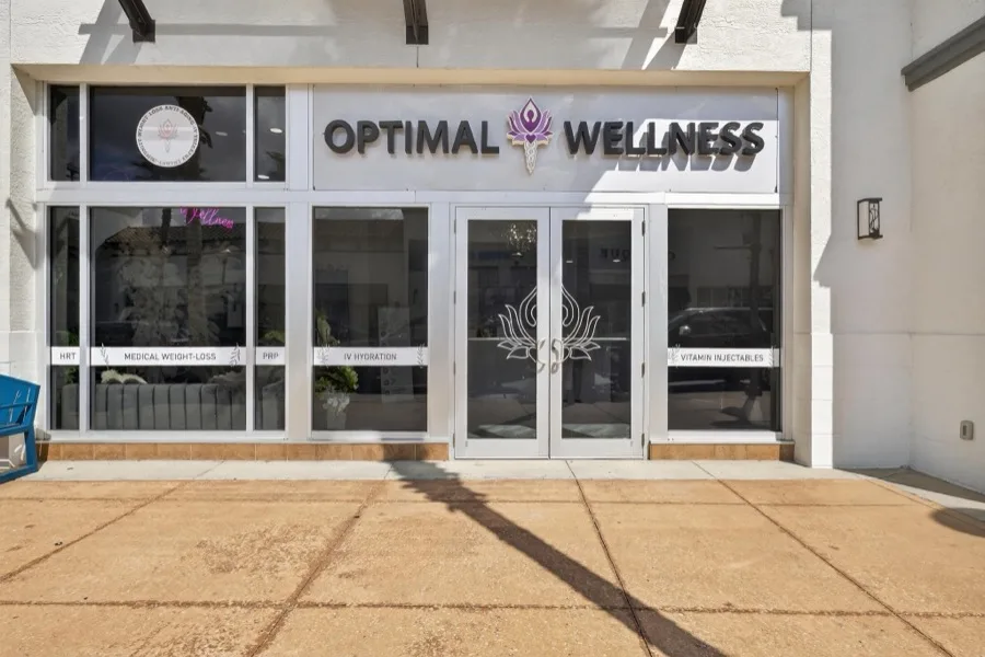 The Optimal Wellness entrance — glass double doors below a black sign carrying the lotus mark, with service decals across the windows