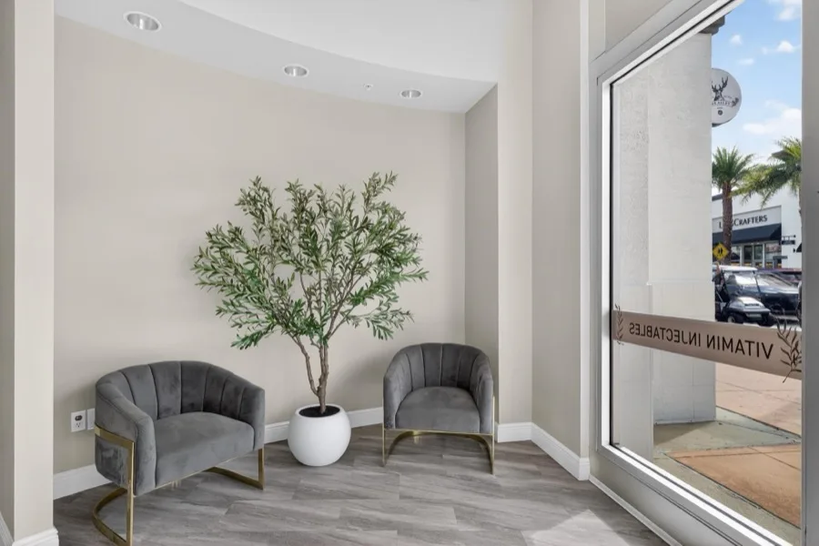 Two grey velvet chairs with gold legs either side of a potted olive tree, beside a window lettered Vitamin Injectables