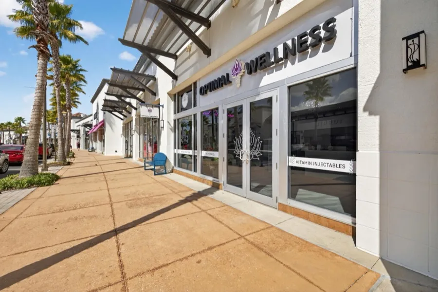 The shopfront seen along the Shops at Wiregrass walkway, palms and blue sky above the Optimal Wellness signage